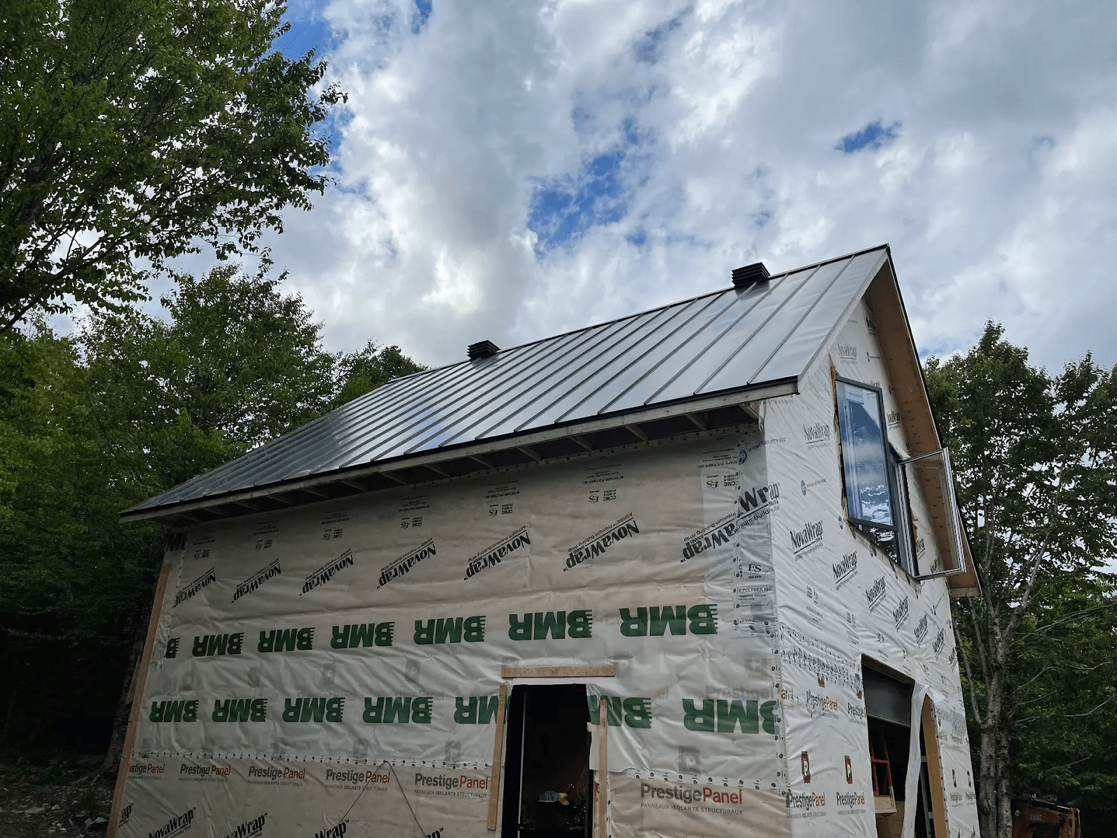 Bâtiment résidentiel en cours de construction, avec un toit en métal gris installé. Les murs sont encore couverts de matériaux de protection et d'isolants temporaires portant les logos 'BMH' et 'PrestigePanel'. Située dans un cadre naturel entouré d'arbres, la structure est conçue avec un toit à pignon et une fenêtre à l'étage. Le ciel nuageux ajoute une atmosphère de chantier en plein air.&nbsp;