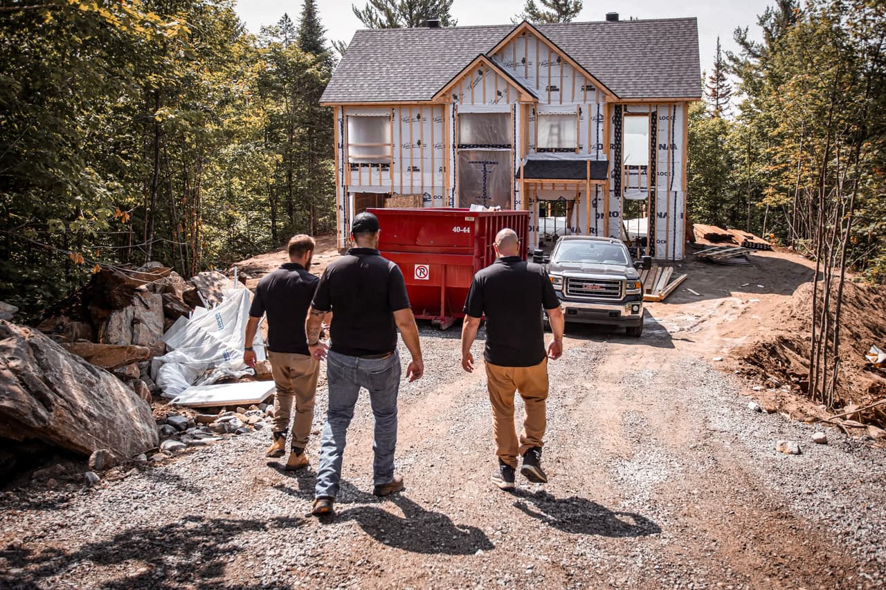 Trois professionnels inspectant un chantier de construction résidentielle avec une maison en construction entourée de végétation.