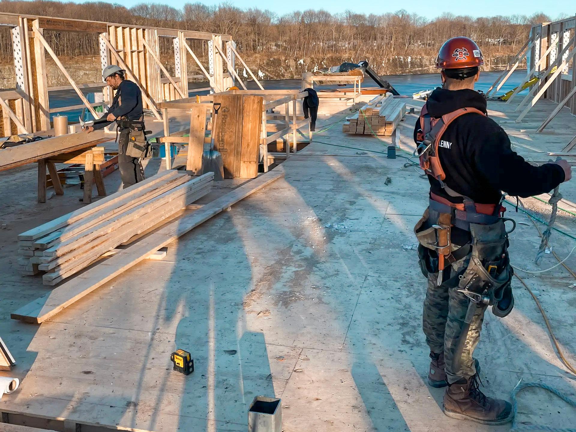 Chantier de construction en bois avec plusieurs ouvriers équipés de harnais de sécurité et casques, manipulant des planches et des matériaux, sous une lumière ensoleillée, dans un environnement ouvert entouré de collines.