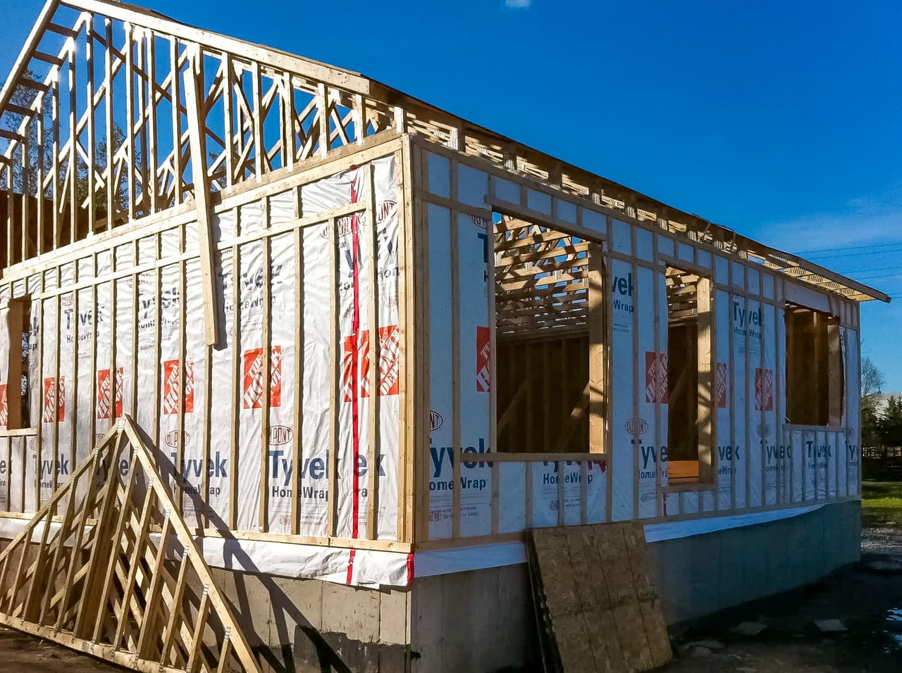 Structure de maison en bois en construction avec pare-intempéries Tyvek, charpente visible et fondations en béton sous un ciel bleu.