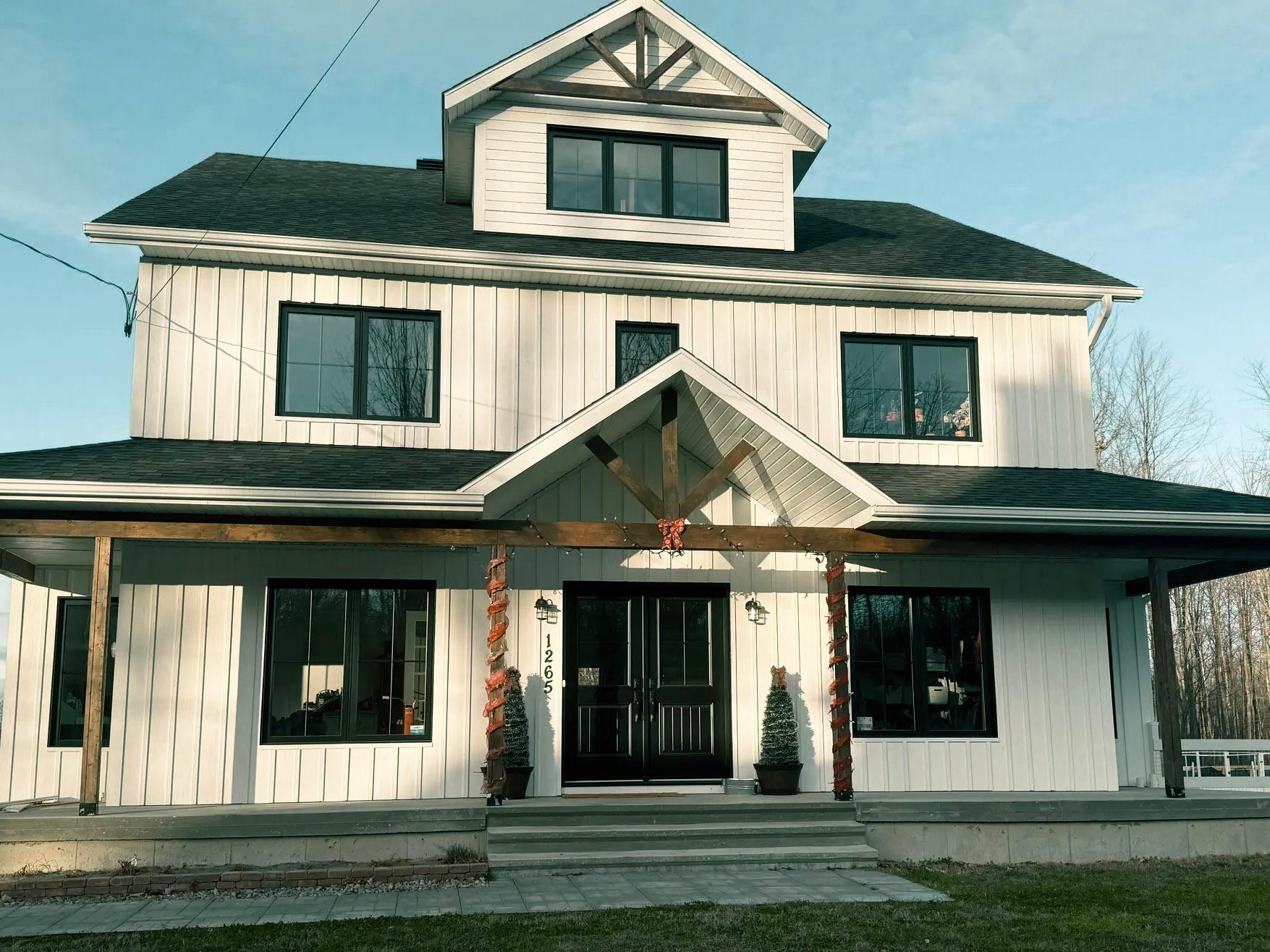 Façade d'une maison moderne à deux étages avec un revêtement extérieur blanc et des accents de bois, dotée d'un porche accueillant décoré de guirlandes oranges pour une ambiance automnale, avec des fenêtres noires et une grande porte d'entrée à double battant.