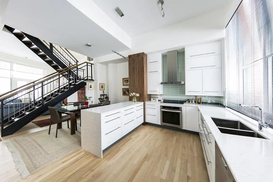 Cuisine contemporaine et lumineuse avec îlot central, plan de travail en quartz blanc et armoires blanches. L'escalier en métal noir ajoute une touche industrielle à cet espace ouvert sur le salon. Parfaite pour les amateurs de lofts et d'espaces ouverts.