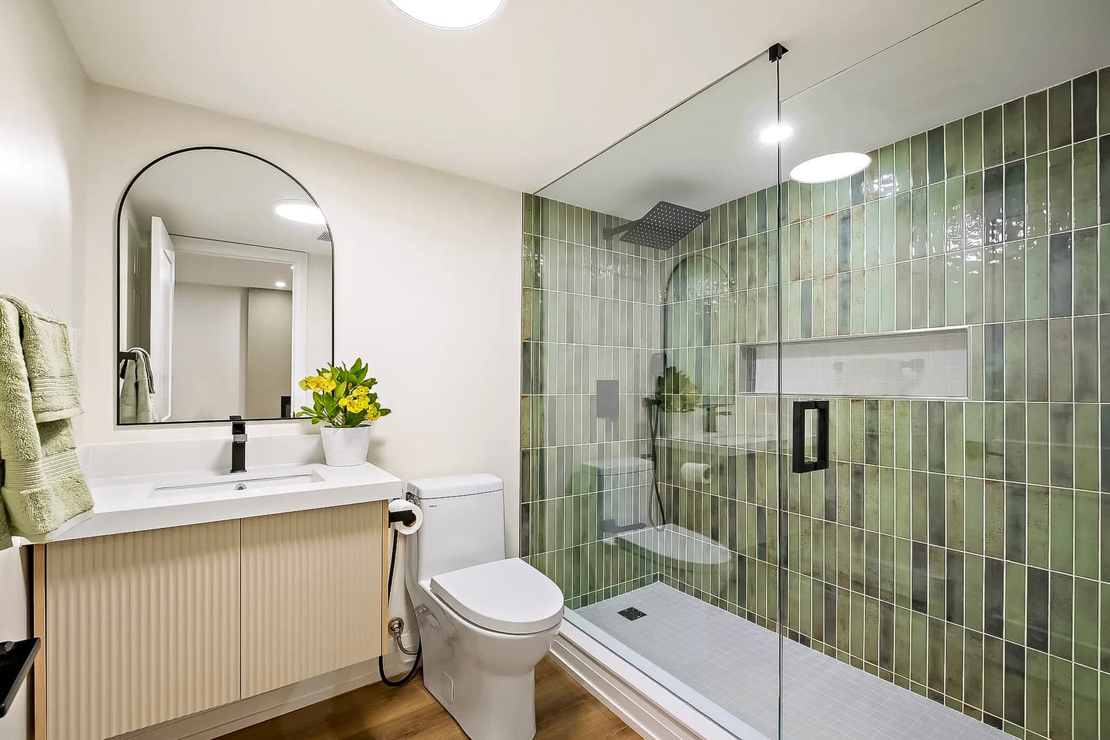 Salle de bain moderne avec douche en verre, carrelage vert, meuble-lavabo en bois clair, miroir arrondi et accessoires élégants.<br>