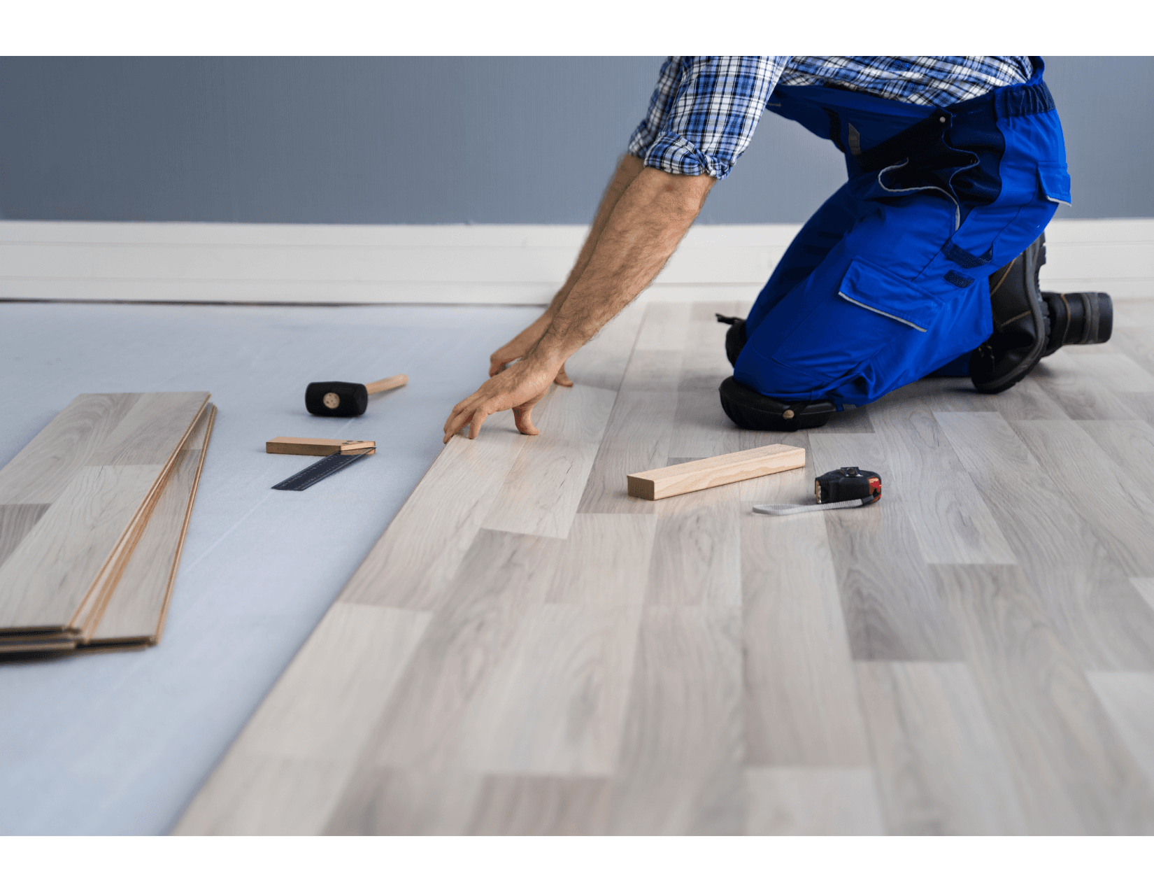 Travailleur posant un nouveau parquet stratifié clair dans une pièce, à genoux sur le sol, entouré d'outils comme un marteau en caoutchouc, une règle et un mètre ruban.