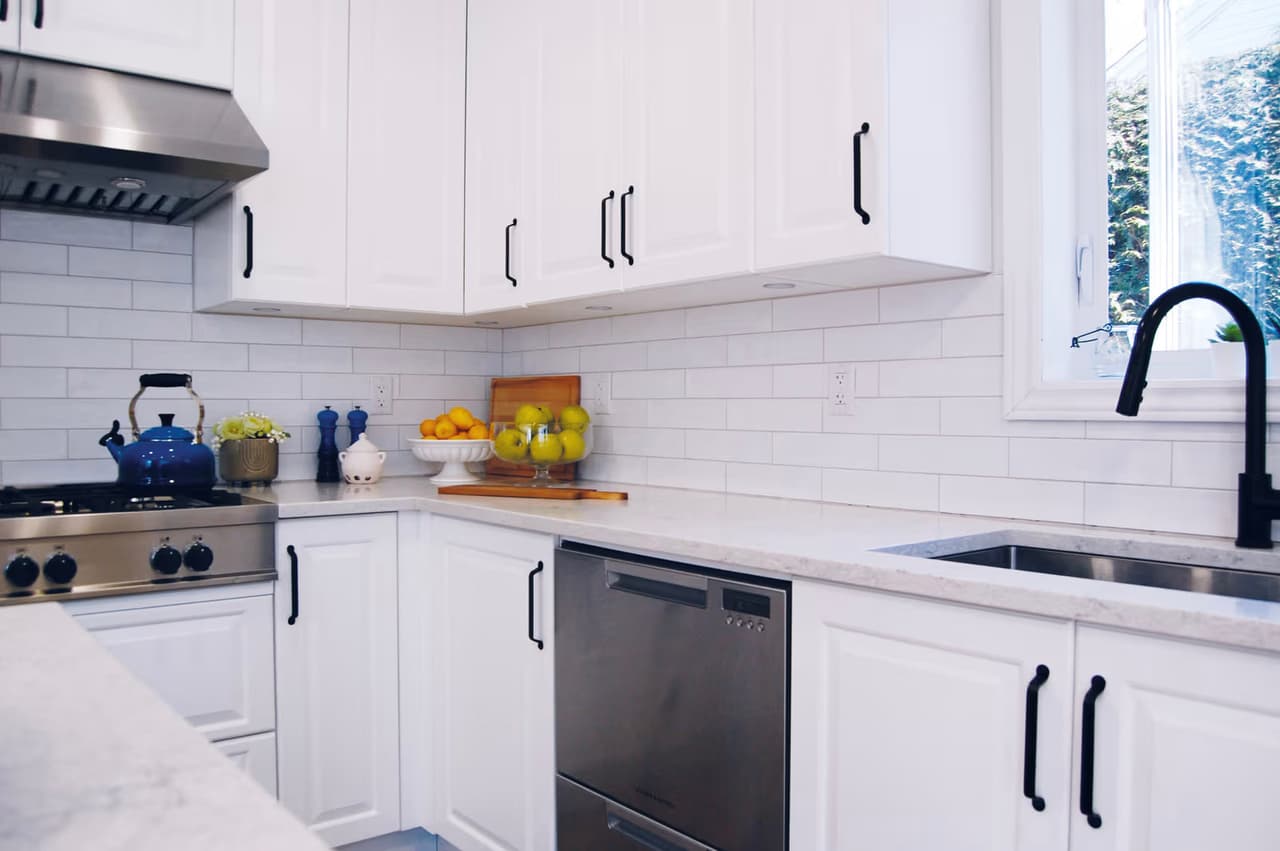 Cuisine moderne blanche avec armoires de style shaker, poignées noires, comptoir clair, dosseret en carreaux métro, électroménagers en acier inoxydable et robinet noir sous la fenêtre.