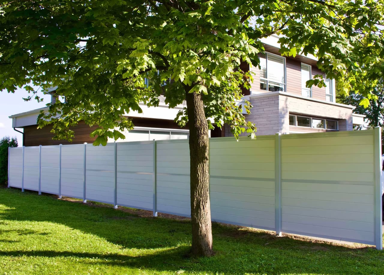 Clôture blanche moderne installée devant une maison contemporaine, entourée d’une pelouse verte et d’un grand arbre mature.