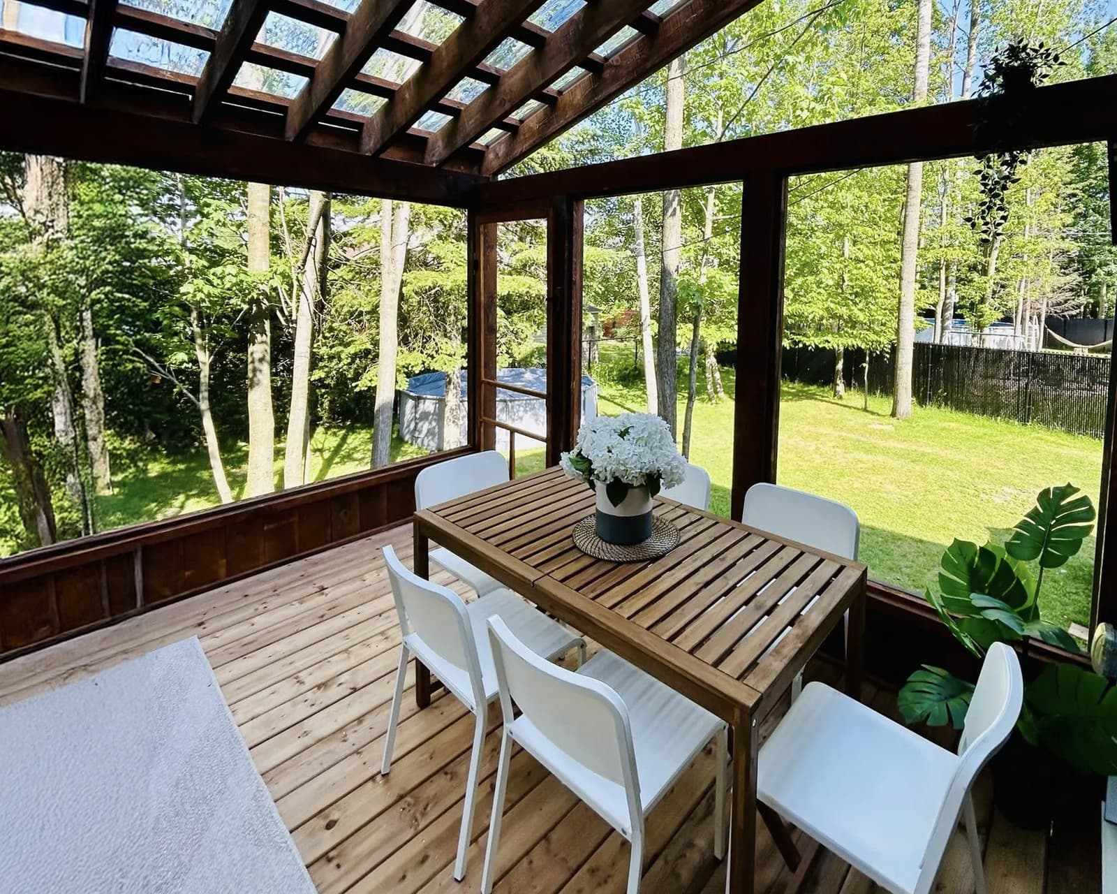 Véranda en bois avec toit vitré, table de jardin en bois et chaises blanches, créant un espace extérieur lumineux et moderne entouré de verdure.