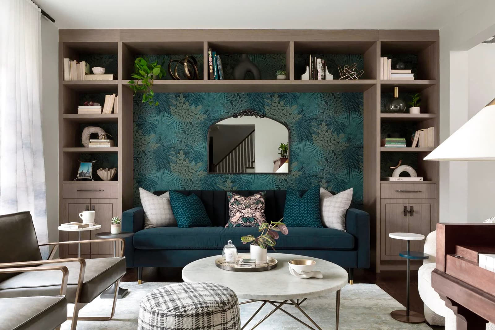 Salon moderne avec sofa en velours bleu, papier peint floral, miroir design, bibliothèque intégrée et table basse en marbre dans un espace lumineux et élégant.