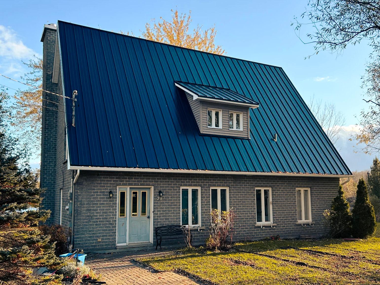 Maison en pierre grise avec un toit en acier bleu foncé à forte pente, dotée d'une lucarne centrale et de grandes fenêtres blanches, entourée d'un paysage automnal avec des arbres et un gazon bien entretenu.