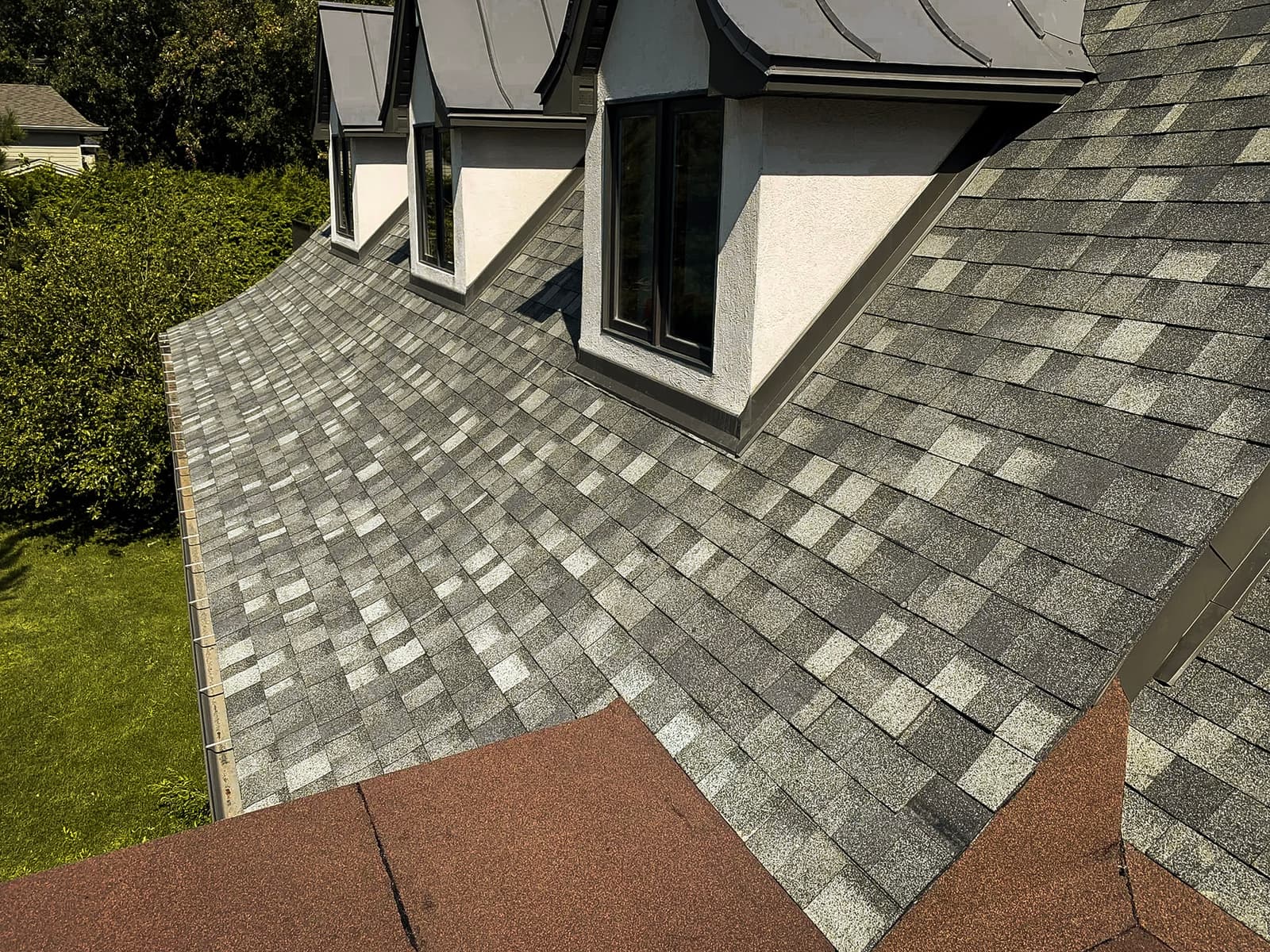 Toit résidentiel avec des bardeaux d'asphalte et des lucarnes offrant une vue sur un jardin verdoyant.<br>