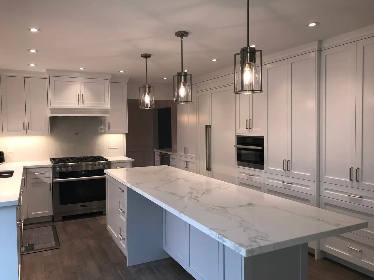 Cuisine moderne blanche avec grand îlot central à comptoir effet marbre, armoires sur mesure, électroménagers encastrés, luminaires suspendus et éclairage encastré.