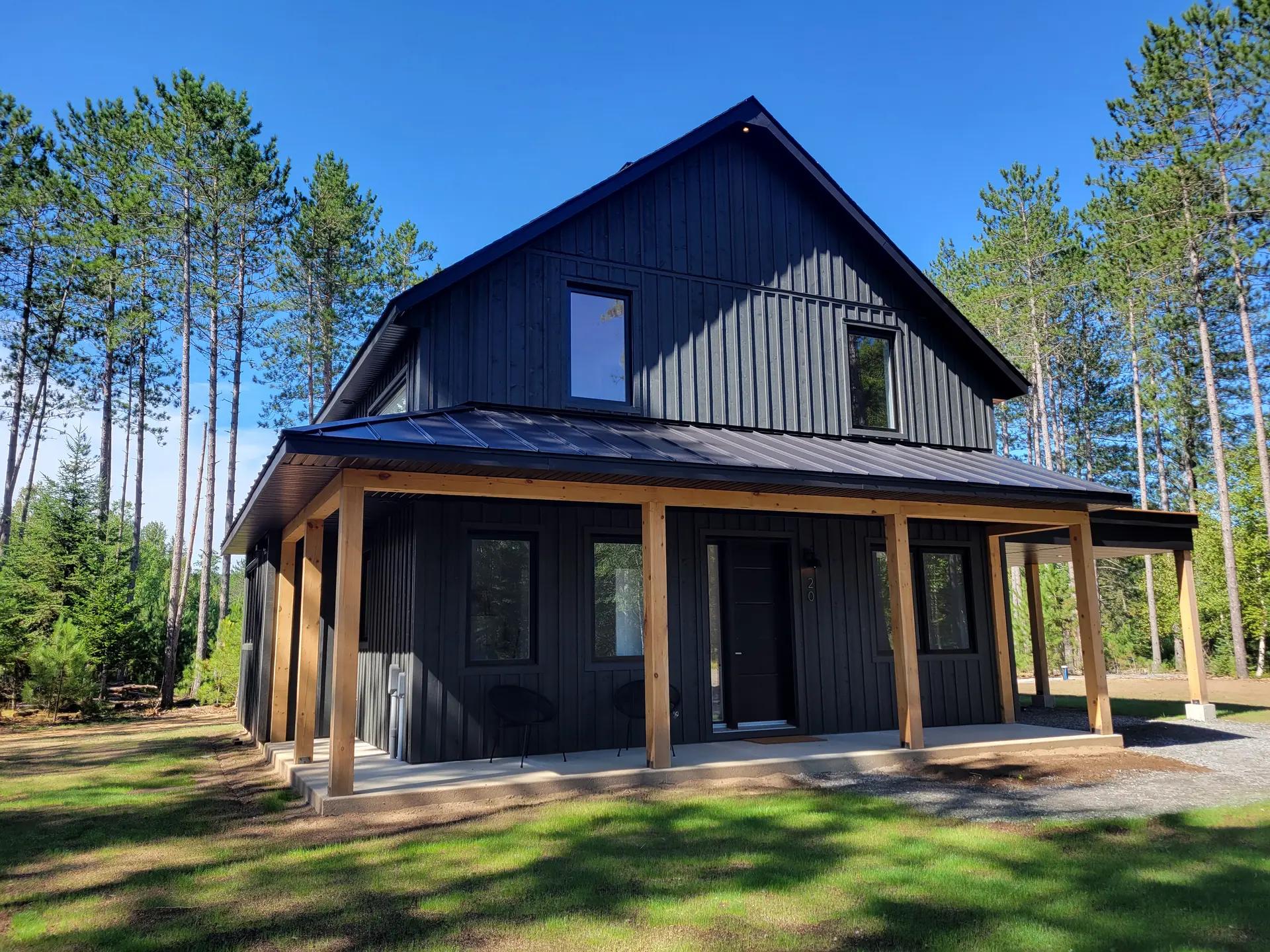 Maison moderne en bois avec un toit en métal, entourée de grands arbres et baignée par le soleil dans un cadre naturel boisé.