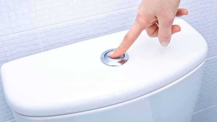 Main appuyant sur le bouton de chasse d’eau d’un réservoir de toilette moderne dans une salle de bain carrelée.