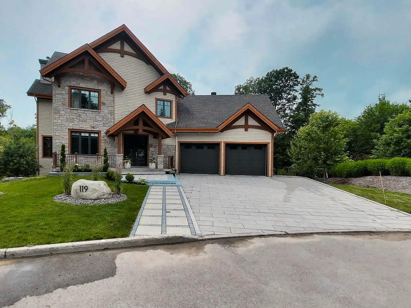 Maison contemporaine en pierre à deux étages avec des accents de bois sur la façade, un grand garage double à portes noires, une allée en pavé moderne et une pelouse bien entretenue, entourée de verdure et d'arbres matures.