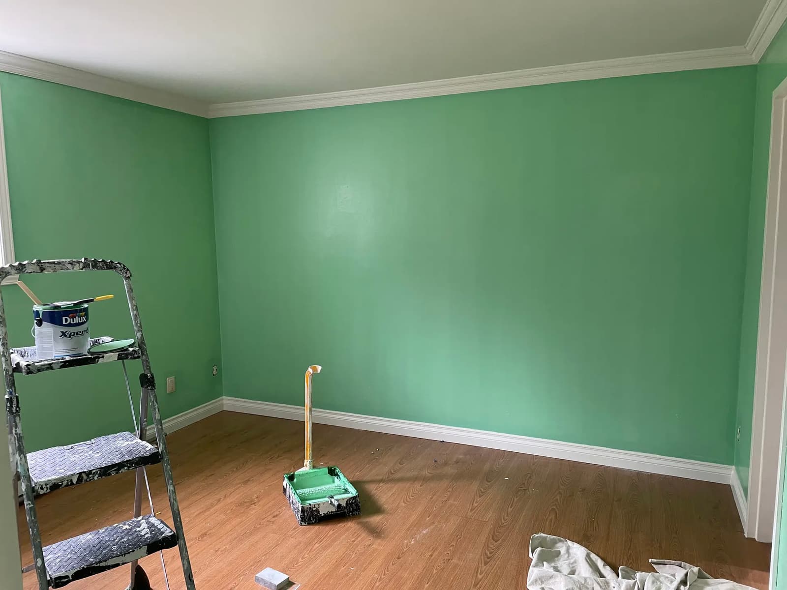 Chambre en cours de rénovation avec murs fraîchement peints en vert, moulures blanches, sol en bois et matériel de peinture au sol.
