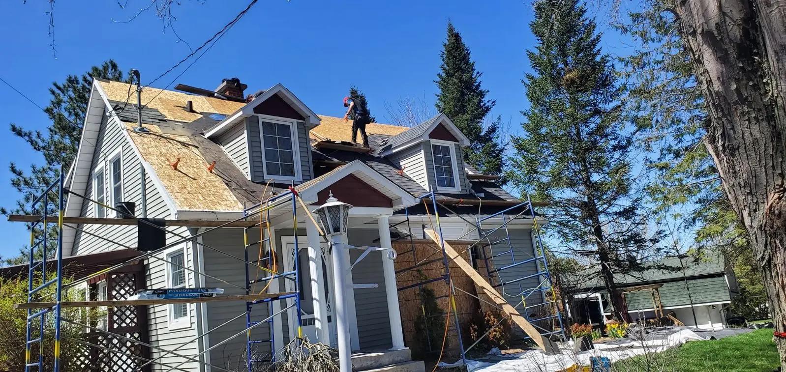 Travaux de rénovation de toiture sur une maison résidentielle à deux étages. Deux ouvriers travaillent sur le toit en installant des panneaux de contreplaqué, entourés d'échafaudages pour assurer leur sécurité. La maison, située dans un cadre verdoyant avec des arbres, présente une façade de couleur grise avec des lucarnes blanches et un porche d'entrée couvert&nbsp;