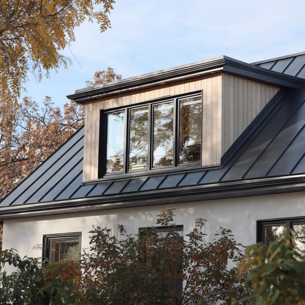 Maison avec toiture métallique noire à joint debout, lucarne moderne, grandes fenêtres et revêtement extérieur clair entouré d’arbres.