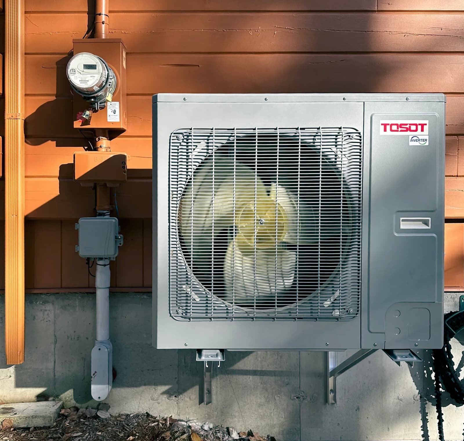 Unité de climatisation extérieure Tosot montée sur un mur en bois orange avec compteur électrique et câblage visible.