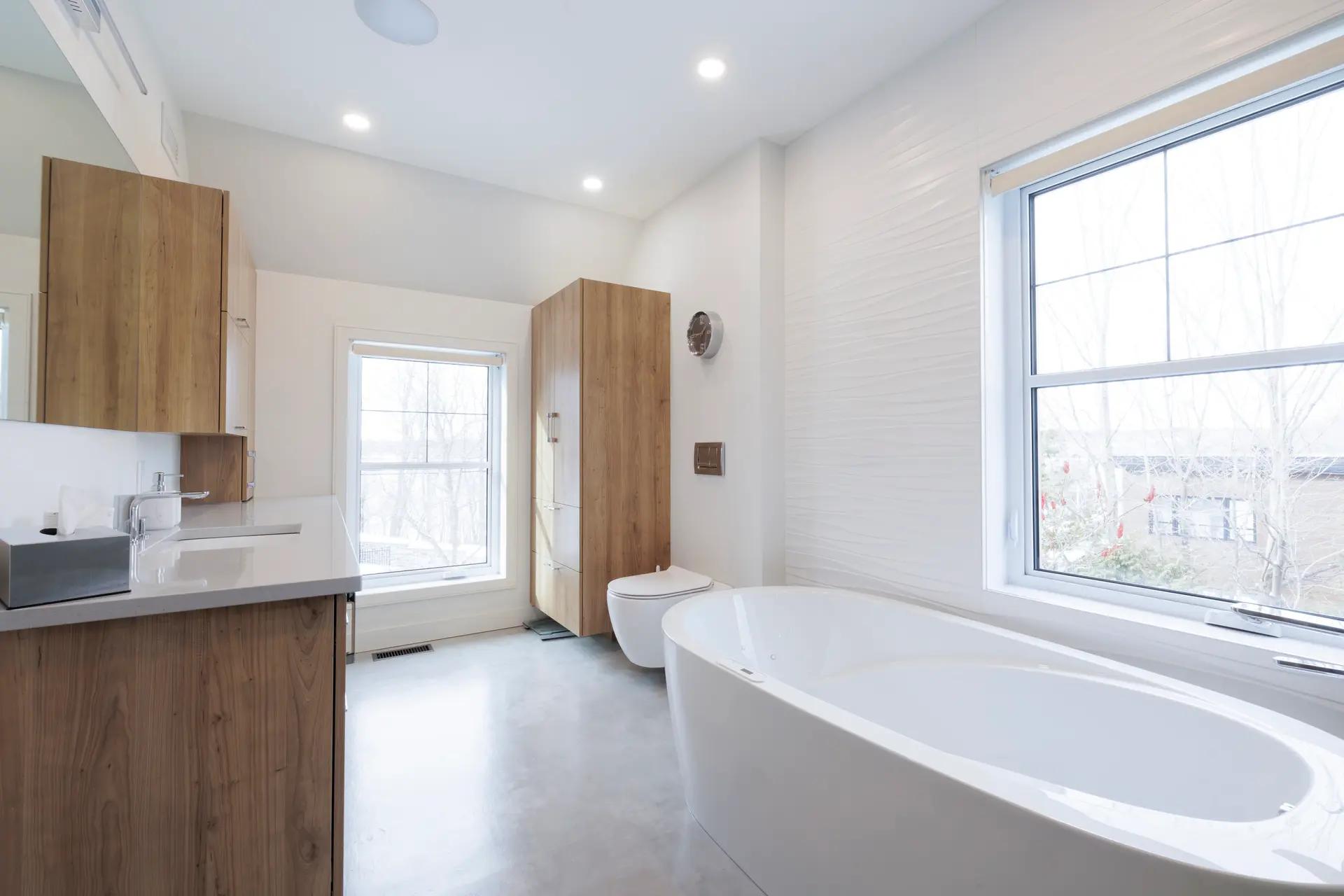 Salle de bains moderne et lumineuse avec baignoire îlot, meuble-lavabo en bois et carrelage blanc.