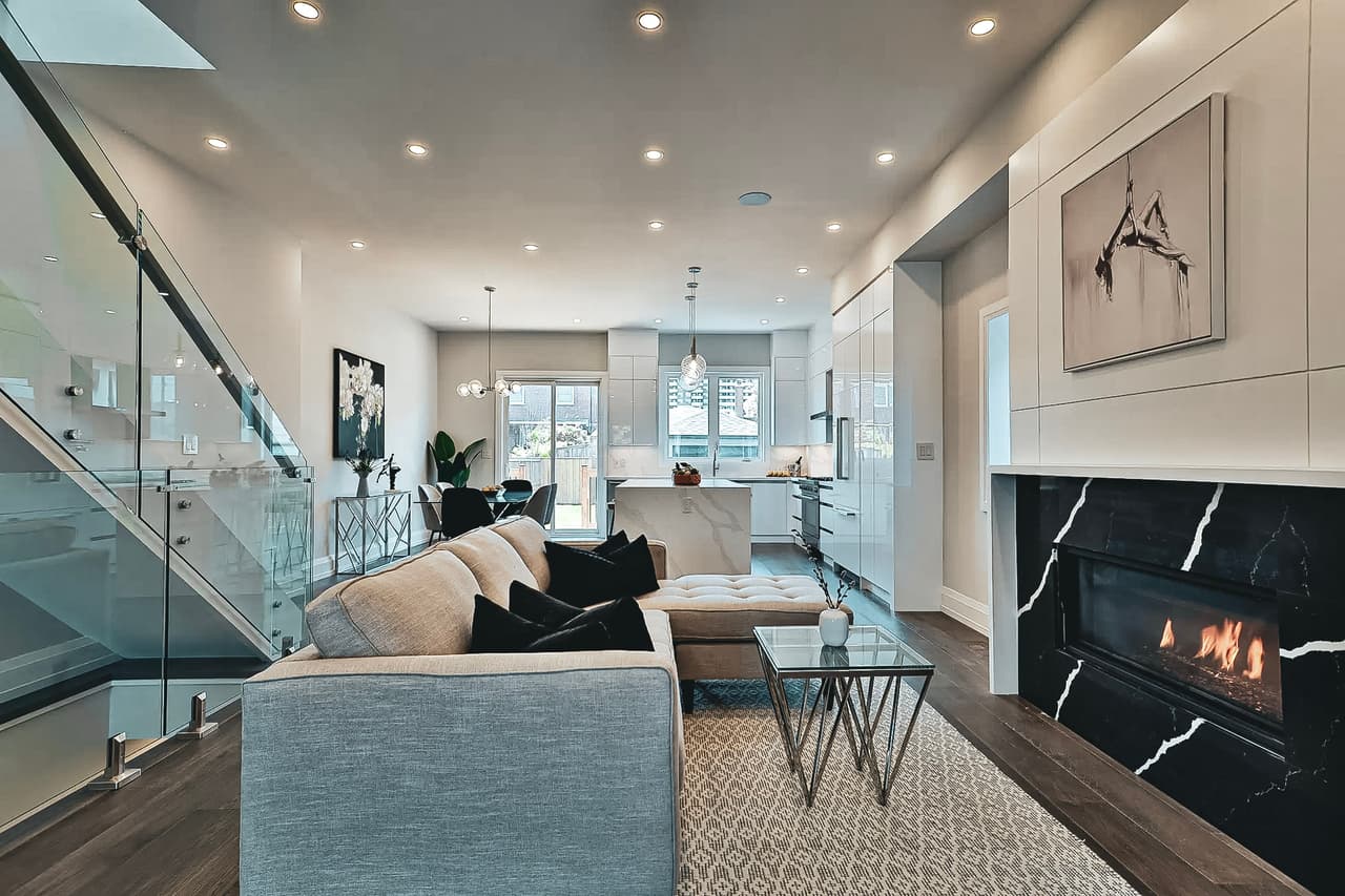 Salon moderne avec foyer en marbre noir, escalier en verre et cuisine ouverte au design épuré.