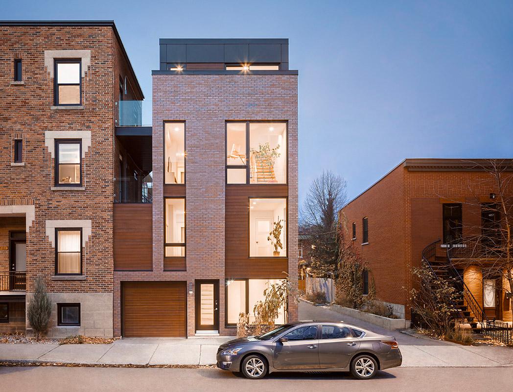 Maison contemporaine située à Montréal, construite en briques rouges avec des panneaux en bois, dotée de grandes fenêtres verticales éclairant l'intérieur, un garage au rez-de-chaussée et un design épuré, entourée de bâtiments urbains similaires dans un quartier résidentiel.