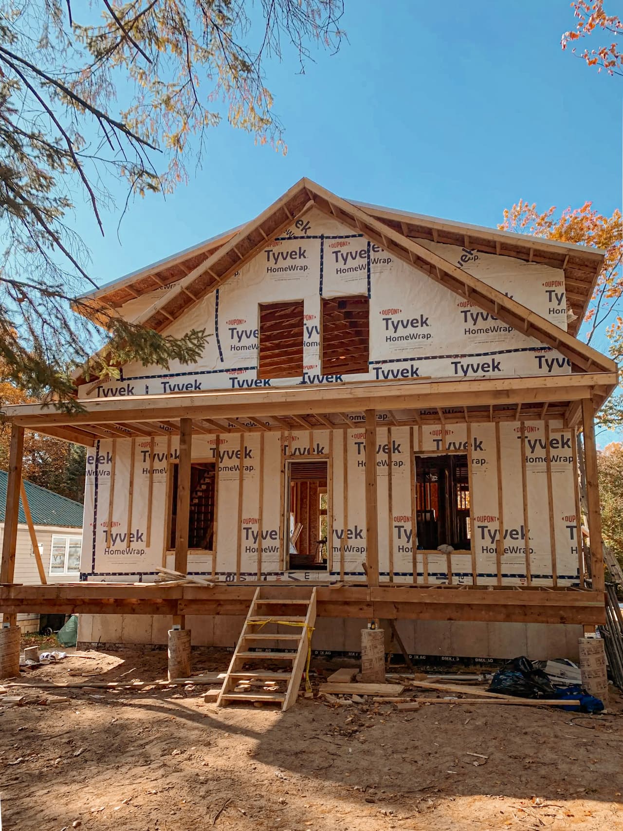 Maison résidentielle en construction avec charpente en bois, membrane pare-air Tyvek, fondation en béton, porche couvert et structure de toiture apparente.