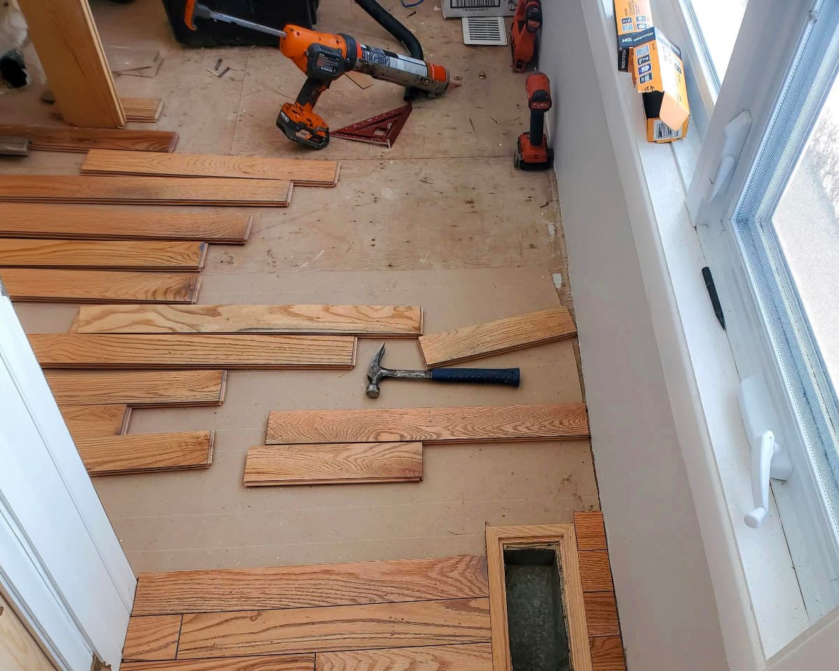 Installation de plancher de bois franc avec outils de construction et lames en cours de pose près d’une fenêtre.