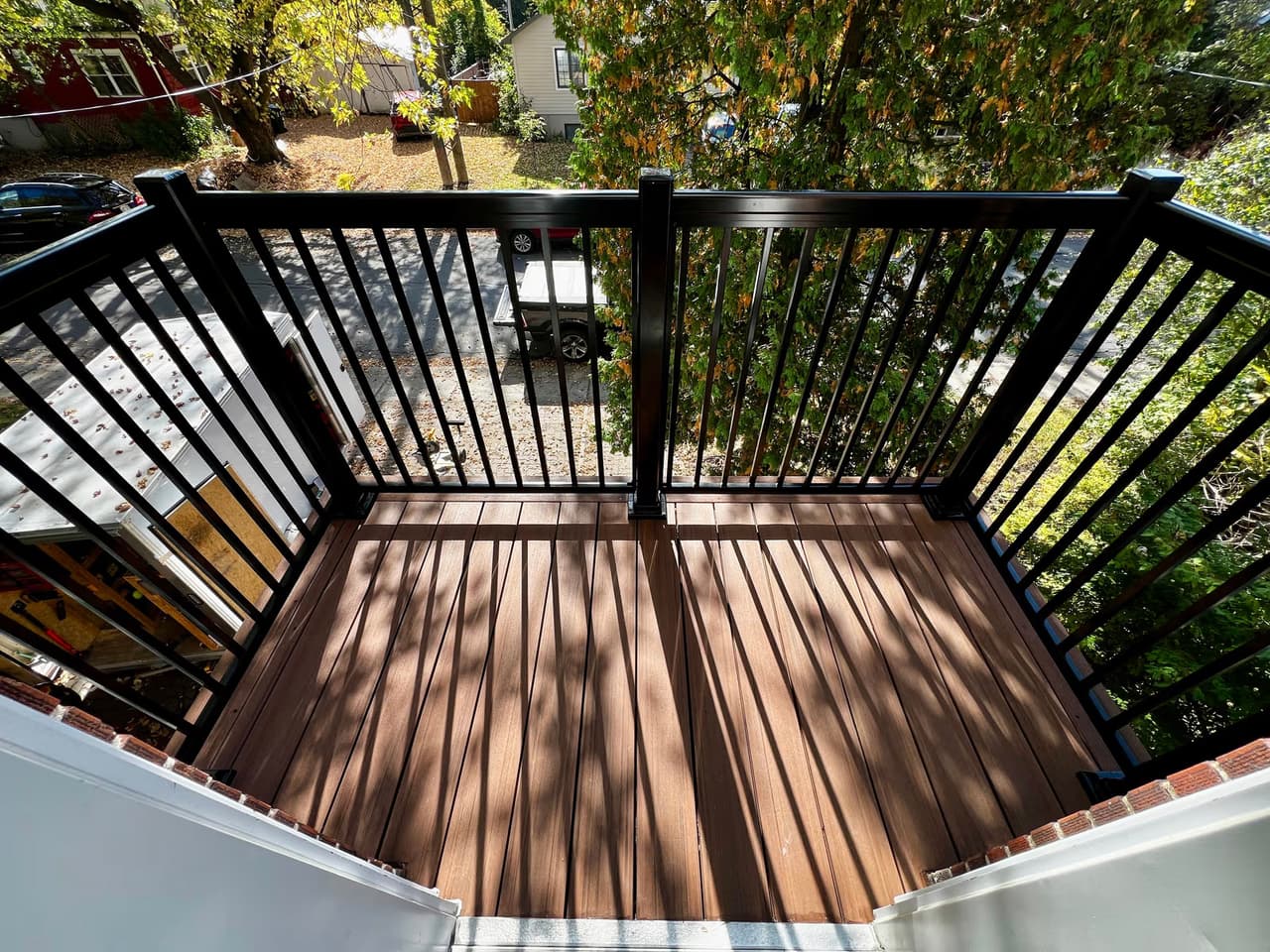 Balcon résidentiel avec plancher en bois brun, garde-corps noir en métal et vue sur arbres, rue et maisons voisines.