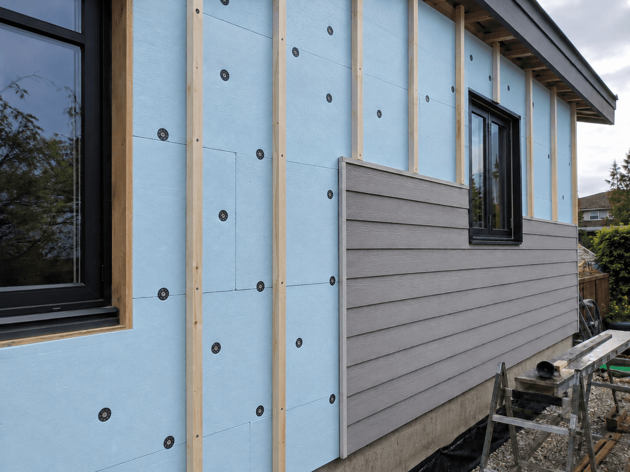 Mur extérieur en rénovation avec panneaux isolants rigides bleus, fourrures de bois, fenêtres noires et revêtement gris partiellement installé