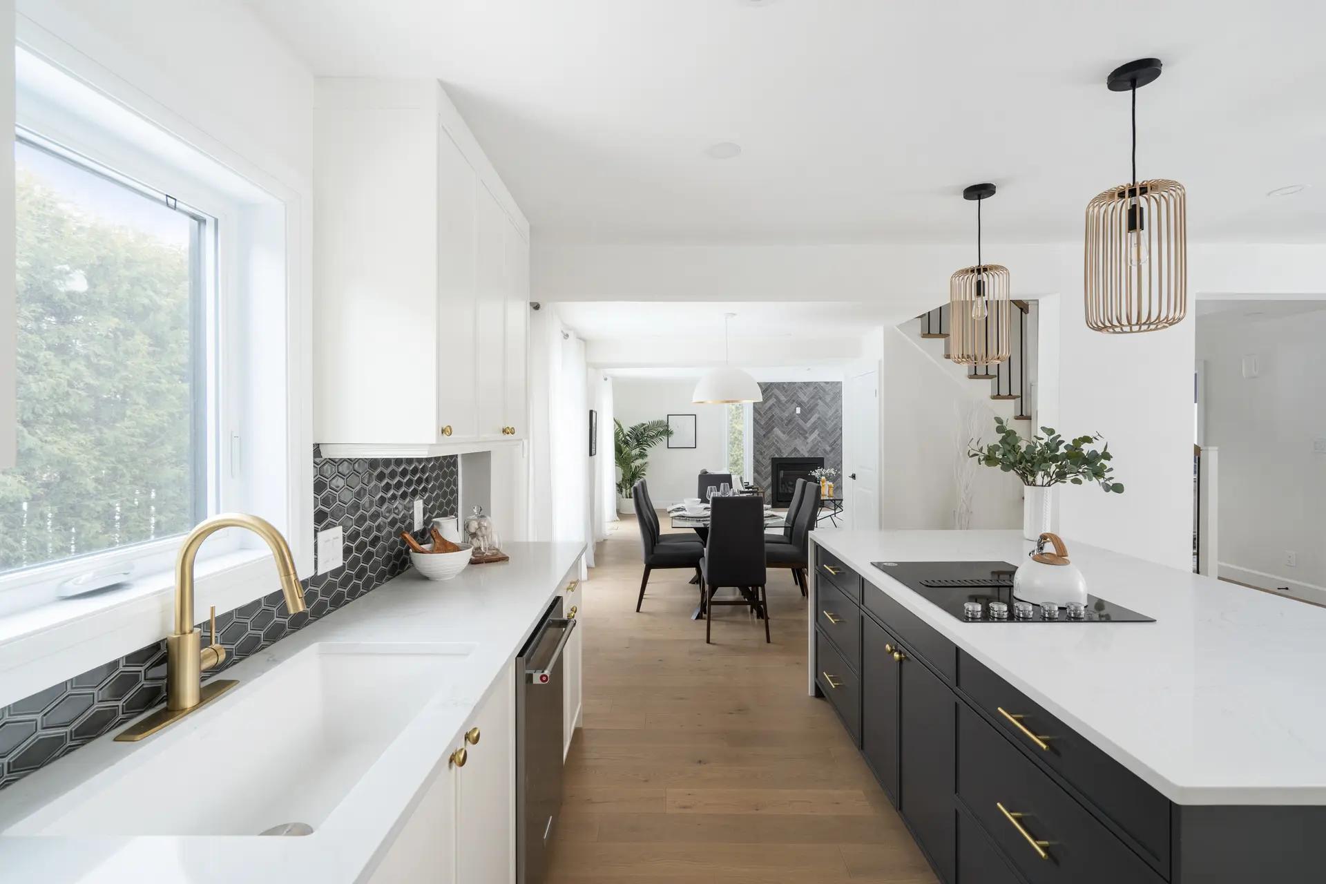 Cuisine de style contemporain à Montréal, mettant en valeur un îlot central noir mat avec un comptoir en quartz blanc. Les armoires blanches shaker et les poignées en laiton apportent une touche d'élégance à l'espace. Le sol en bois clair et les luminaires suspendus créent une ambiance chaleureuse et accueillante