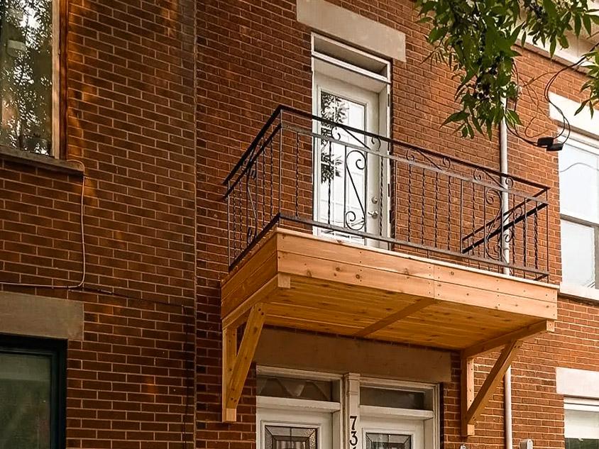 Petit balcon en bois avec garde-corps en fer forgé noir orné de motifs décoratifs, situé à l'étage d'une maison en briques rouges avec une porte blanche donnant accès à l'extérieur, entouré d'un cadre urbain avec des arbres en arrière-plan.