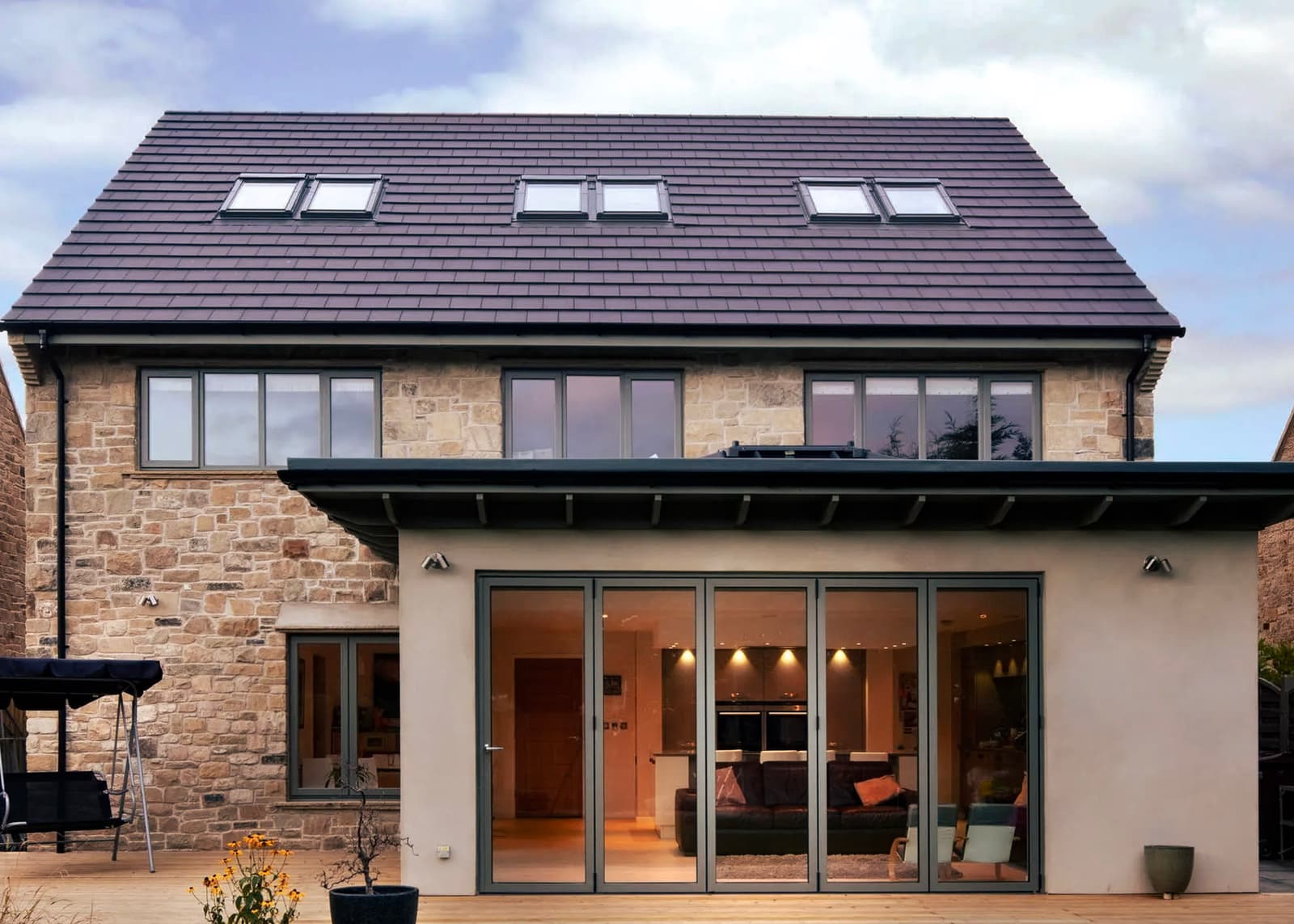 Maison en pierre rénovée avec toit en tuiles, fenêtres à double vitrage, portes-fenêtres pliantes, terrasse en bois et éclairage extérieur moderne.