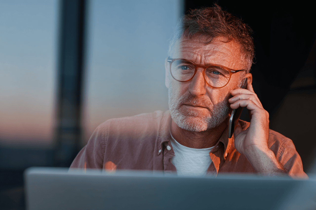 Homme concentré avec des lunettes parlant au téléphone devant un ordinateur portable, en fin de journée.