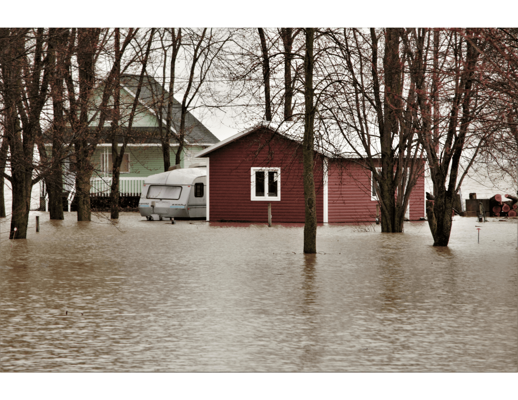 Inondation dans une zone résidentielle, avec une maison rouge entourée d'eau et un camping-car stationné à proximité. Des arbres dénudés par l'hiver se dressent au milieu des eaux stagnantes.