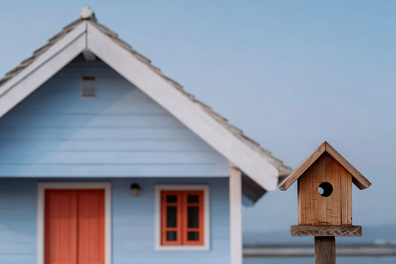 Nichoir en bois fixé sur un poteau devant une maison bleue avec porte et fenêtre rouges, toit en pente et arrière-plan extérieur légèrement flou.