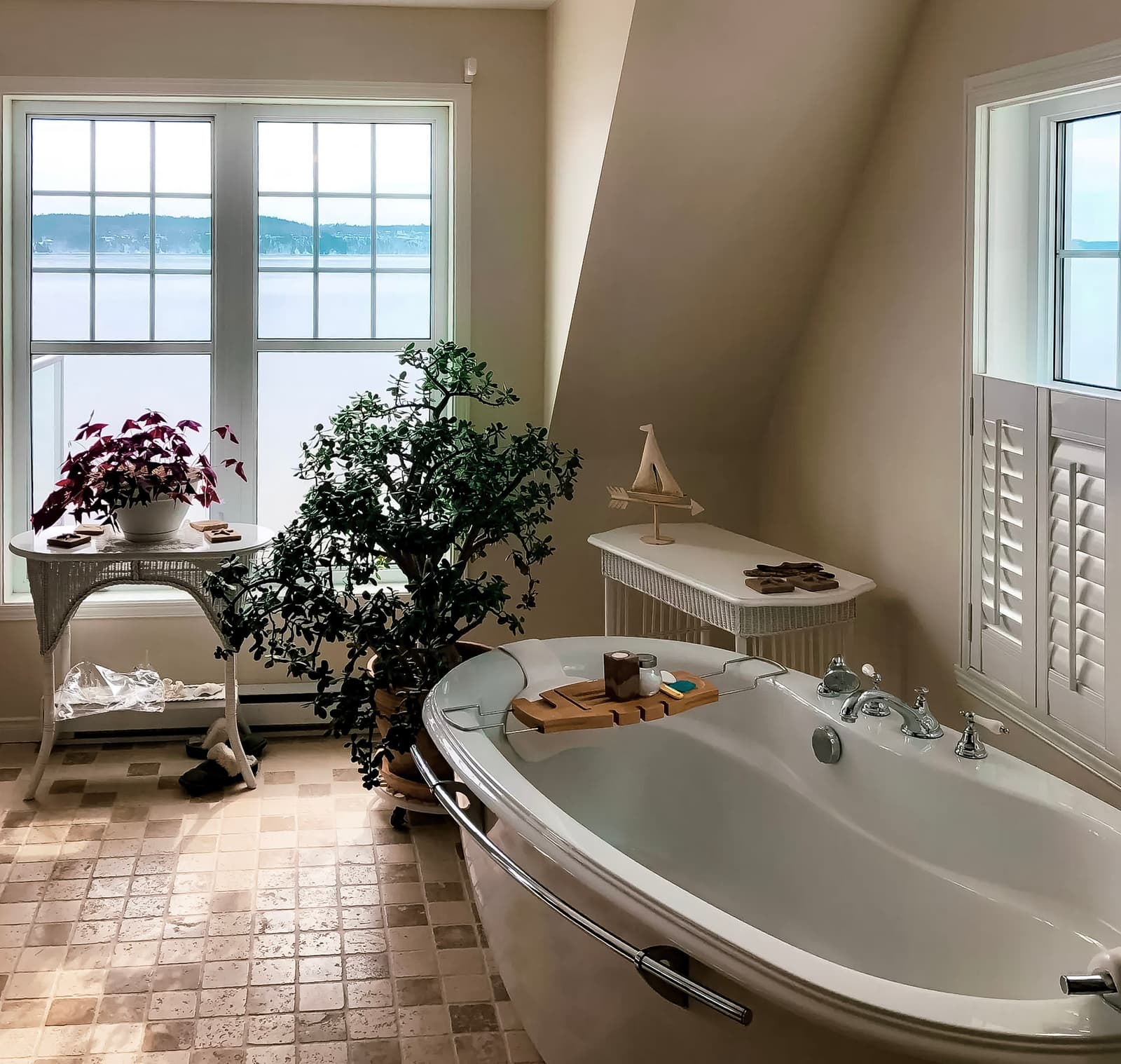 Salle de bain lumineuse avec baignoire autoportante, carrelage en pierre naturelle et grandes fenêtres avec vue sur un paysage extérieur. Mobilier en rotin blanc et décoration végétale apaisante.