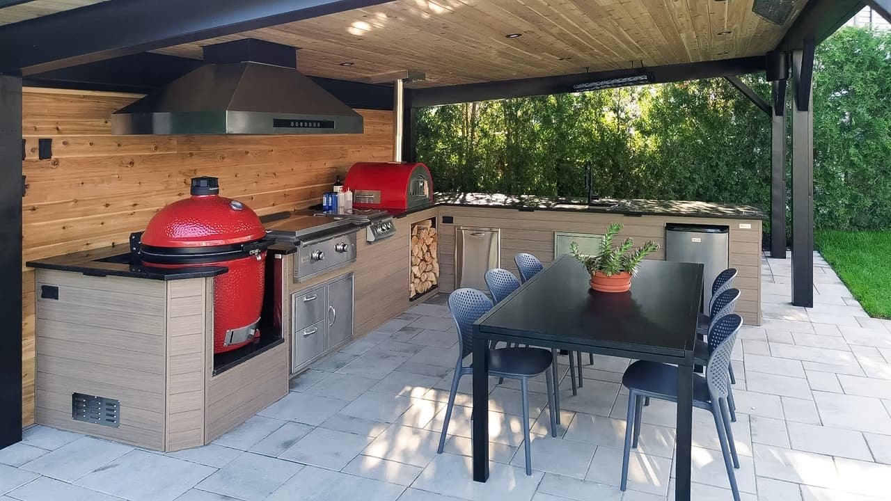 Cuisine extérieure couverte avec barbecue, fumoir, four à pizza, hotte, comptoirs, rangements intégrés et salle à manger sur patio pavé.