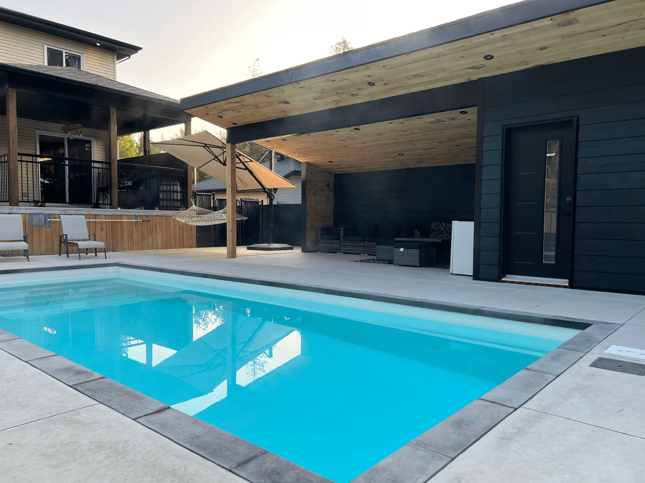 Cour arrière moderne avec piscine creusée, terrasse en dalle de béton, pavillon noir avec plafond en bois, éclairage encastré, porte extérieure et espace détente couvert