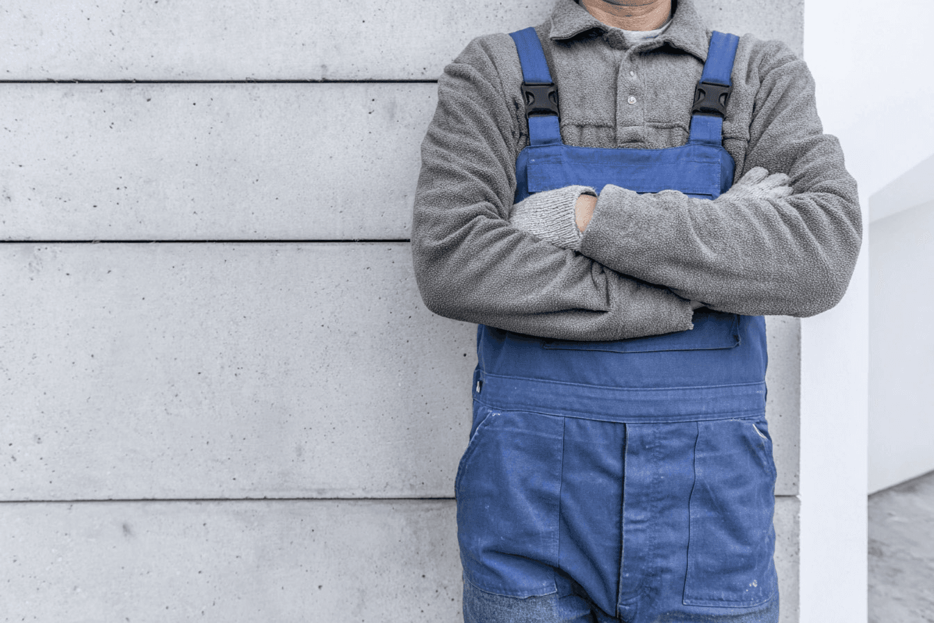 Plombier professionnel en bleu de travail et gants de chantier, debout les bras croisés devant un mur en béton.

