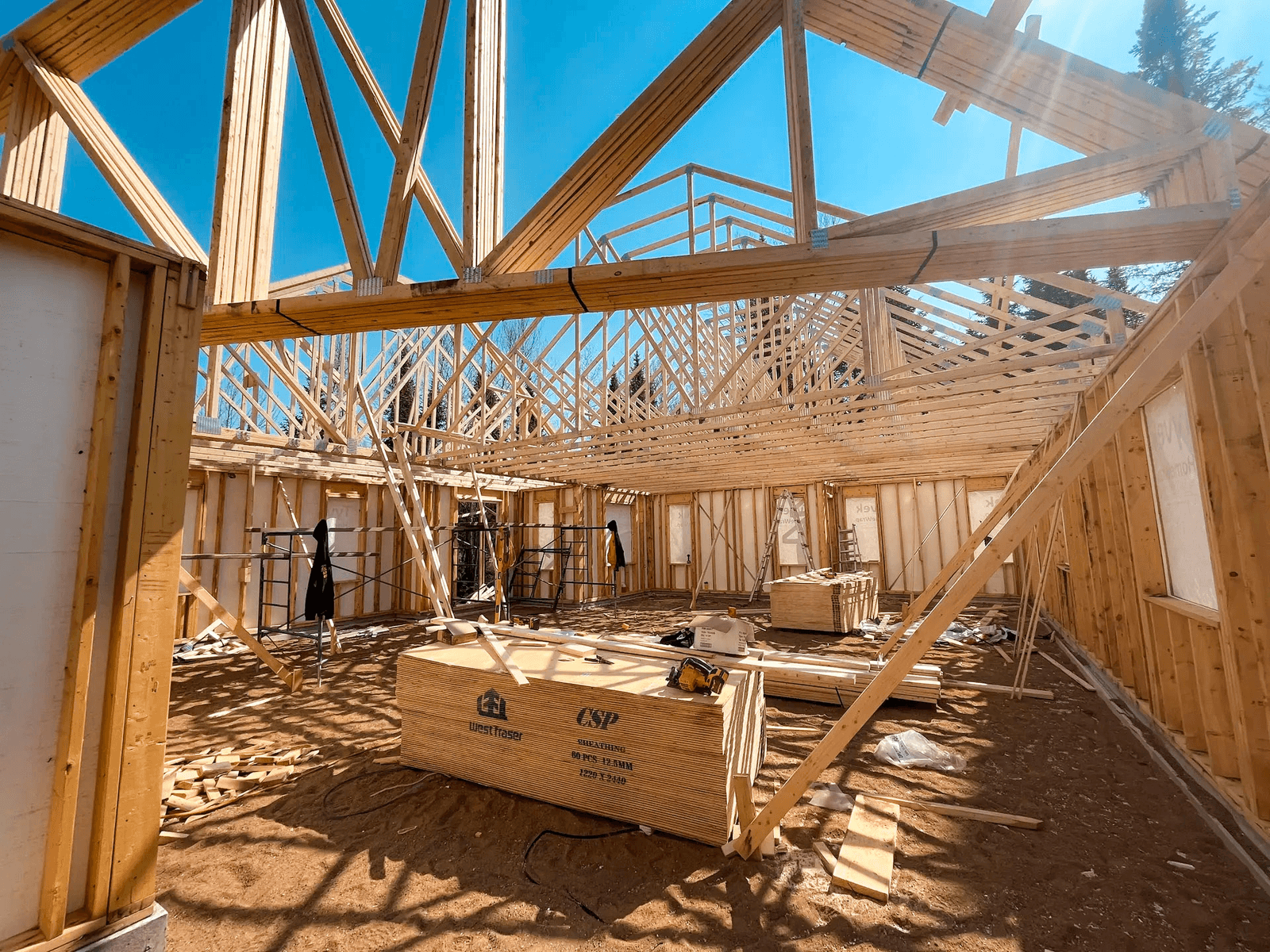 Intérieur d’une maison en construction avec charpente en bois apparente, fermes de toit, murs à ossature, matériaux de construction et équipements de chantier.