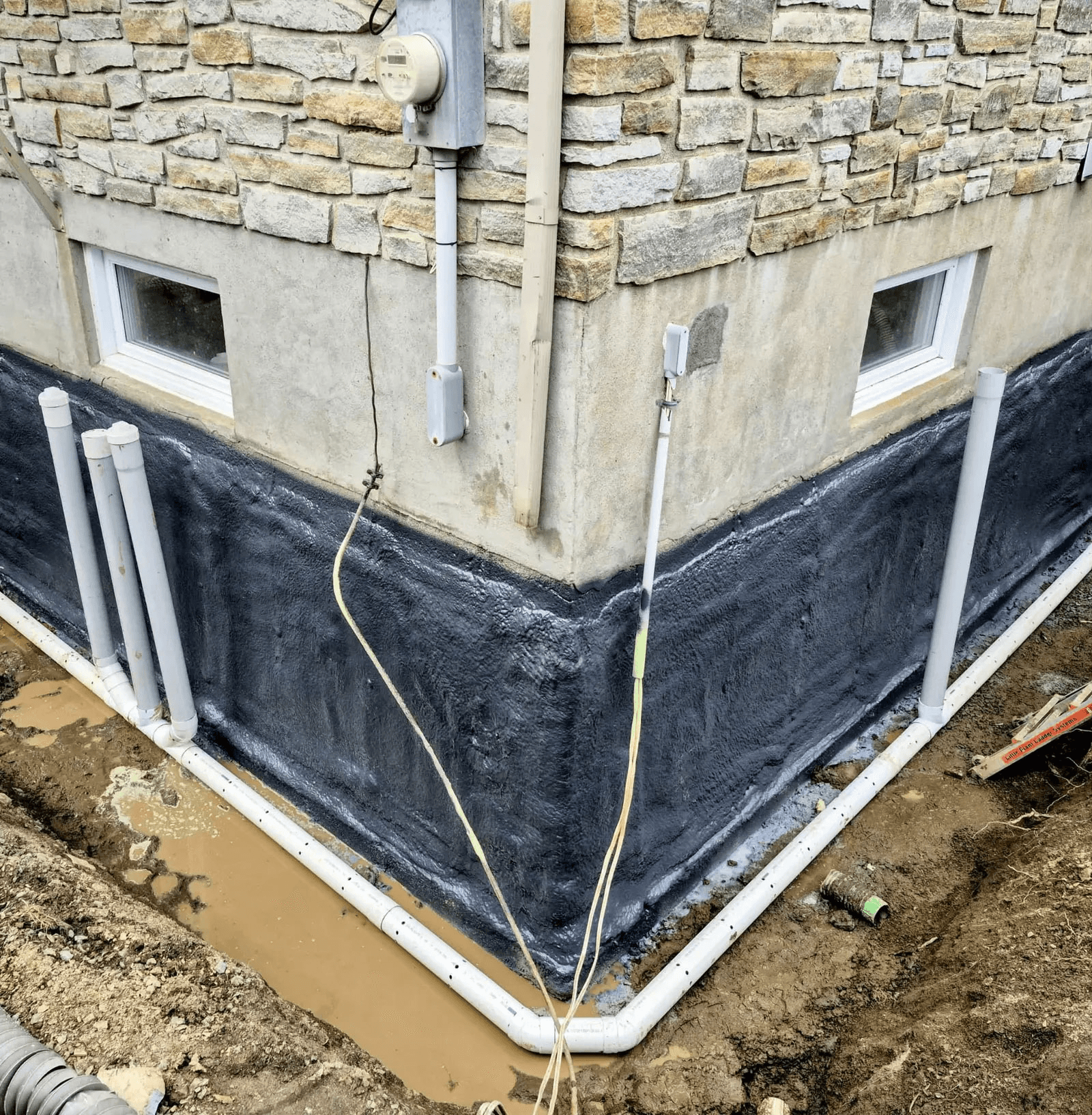 Installation d’un drain français en PVC autour des fondations d’une maison avec application d’un enduit imperméabilisant noir pour prévenir les infiltrations d’eau.