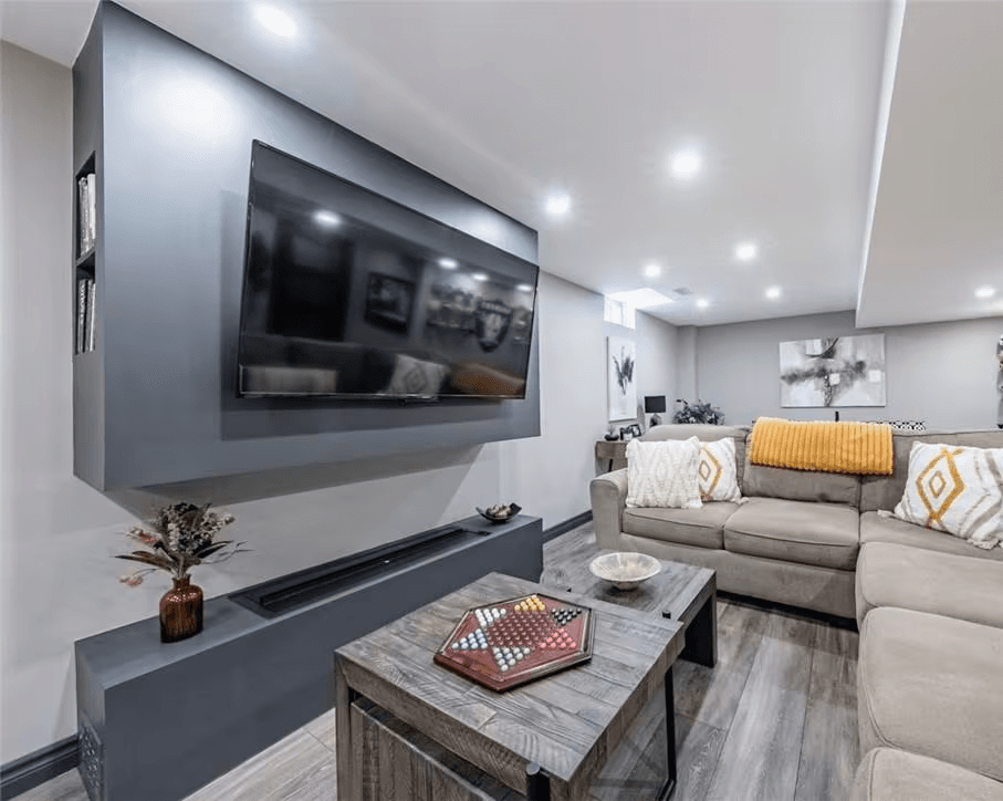 Salle familiale moderne au sous-sol avec grand téléviseur mural, canapé sectionnel gris, tables en bois, éclairage encastré et plancher effet bois