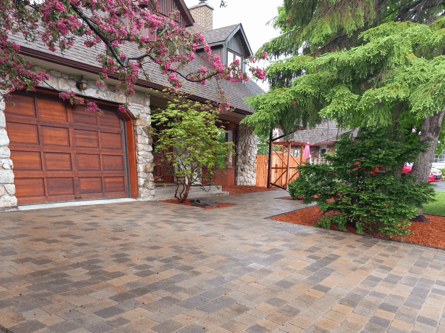 Entrée résidentielle en pavé uni multicolore devant une maison en pierre et bois, avec garage en bois, arbres matures et aménagement paysager soigné