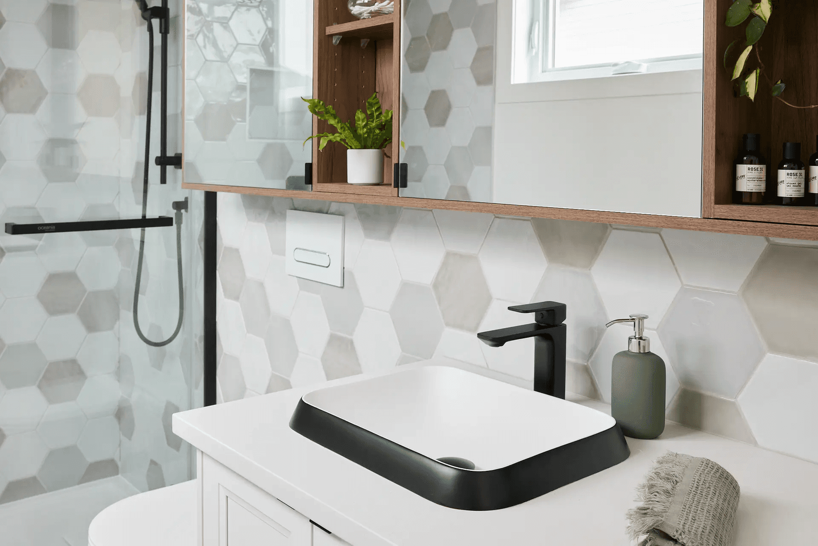 Salle de bain moderne avec carrelage mural hexagonal blanc, gris et beige, douche vitrée à robinetterie noire, lavabo vasque rectangulaire blanc et noir, comptoir blanc, meuble-lavabo et armoires murales en bois avec rangements décoratifs