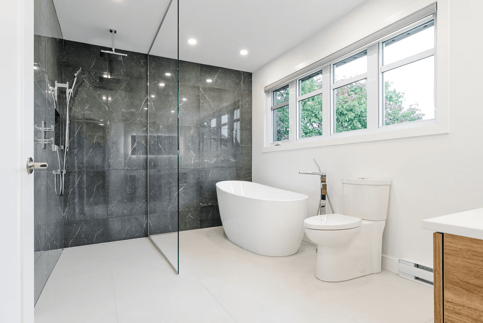 Salle de bain moderne avec douche à l’italienne vitrée, grand carrelage anthracite effet pierre, baignoire autoportante blanche et toilette sous une large fenêtre&nbsp;