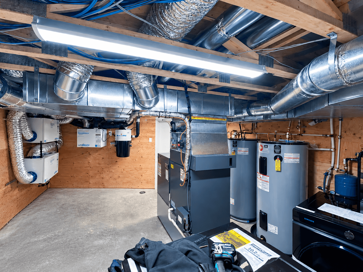 Salle mécanique résidentielle équipée d’un système de chauffage et ventilation, conduits métalliques, chauffe-eau, échangeurs d’air, tuyauterie apparente et équipements techniques.