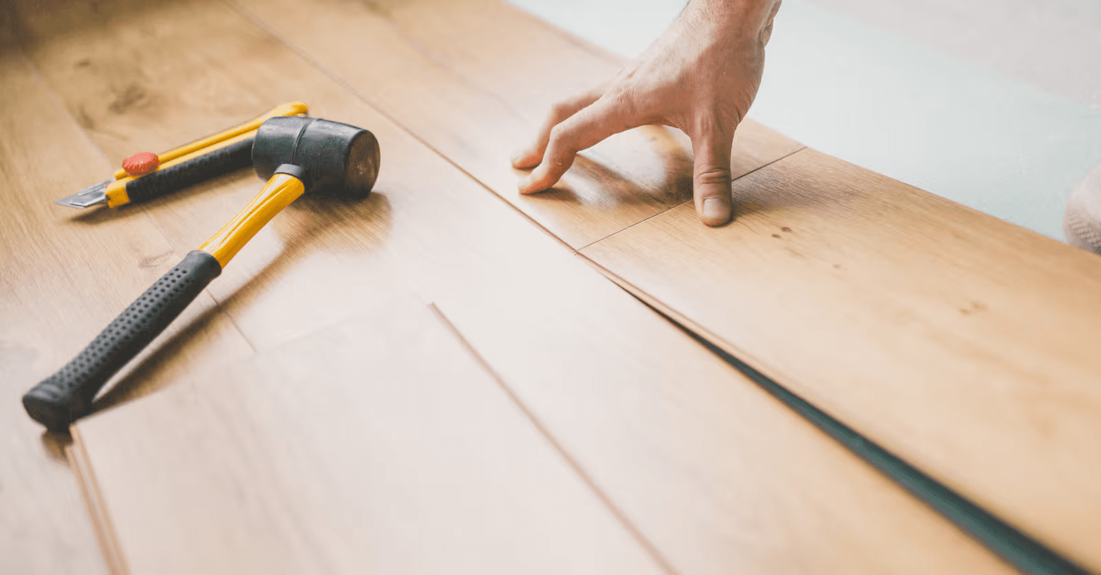 Installation d’un plancher flottant en lames de bois clair par un professionnel ajustant les joints des planches à la main, avec maillet en caoutchouc et couteau utilitaire, lors de travaux de rénovation intérieure