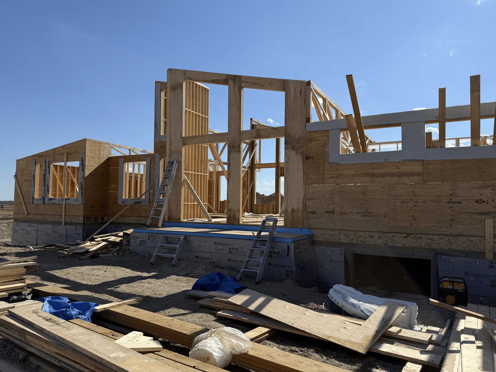 Maison en construction avec ossature en bois, murs structuraux, ouvertures pour portes et fenêtres, fondation isolée et matériaux de chantier.