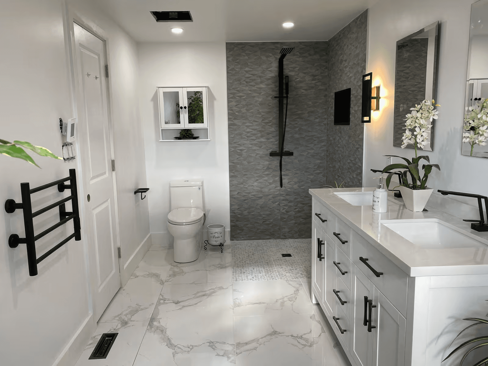 Salle de bain moderne rénovée avec douche à l’italienne en carreaux gris, double meuble-lavabo blanc, robinetterie noire et plancher effet marbre