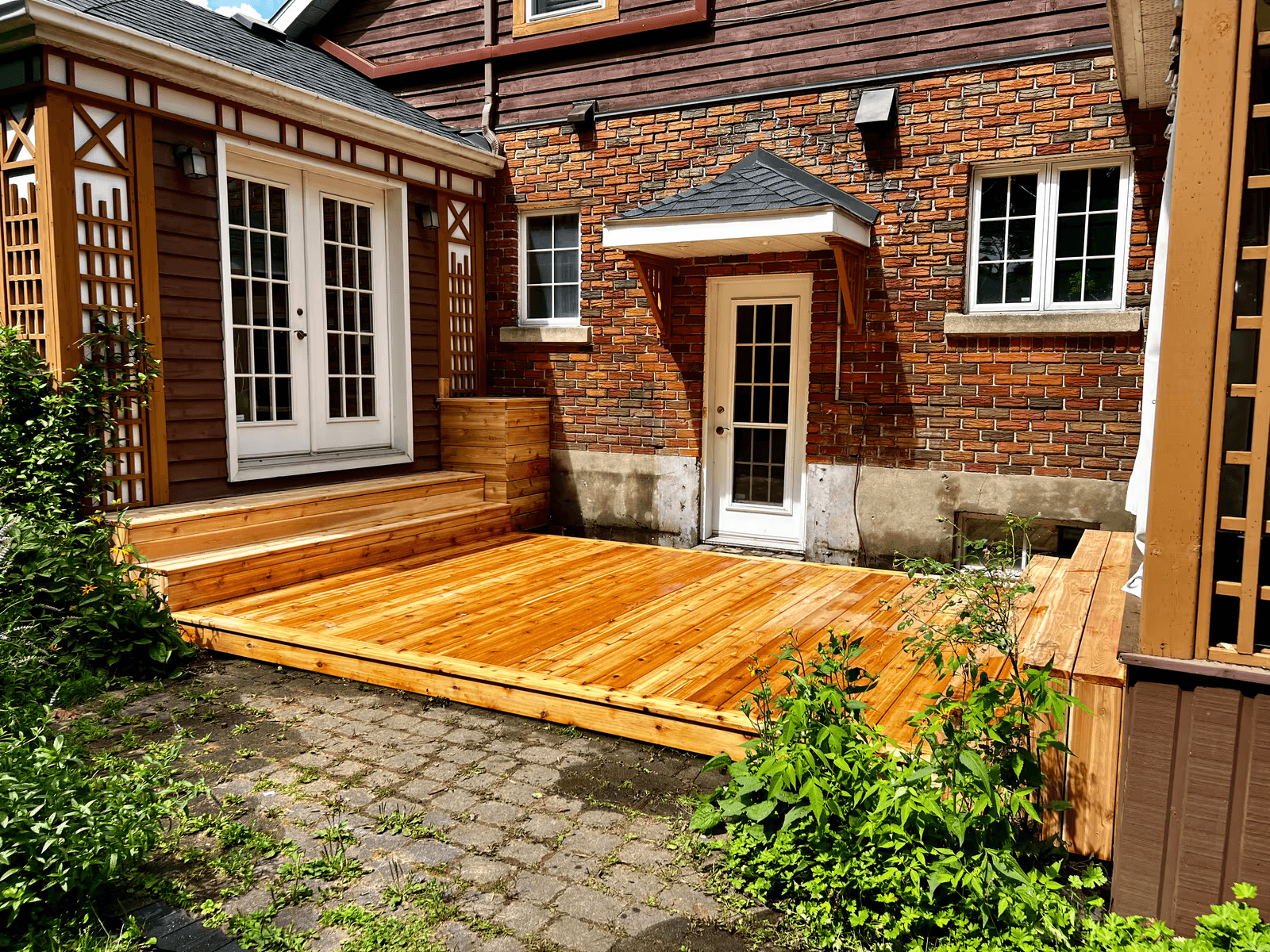 Terrasse extérieure résidentielle en bois naturel avec plancher neuf, marches larges, portes françaises blanches, treillis décoratifs en bois et aménagement paysager, aménagée dans une cour intérieure bordée de murs en brique rouge et de revêtement brun